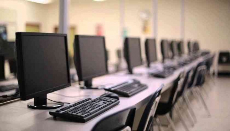 Ruangan yang remang-remang memiliki papan nama bercahaya mencolok yang menampilkan kata-kata 'Sysco LABS.' Di bawah papan nama tersebut terdapat meja kantor dengan berbagai perangkat elektronik, termasuk laptop dan kabel. Pencahayaan tersebut menciptakan suasana yang sejuk dan profesional.